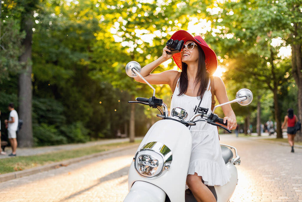 attractive woman riding on motorbike in street, summer vacation style, traveling, smiling, happy, having fun, stylish outfit, adventures, taking pictures on vintage photo camera - Photo, Image
