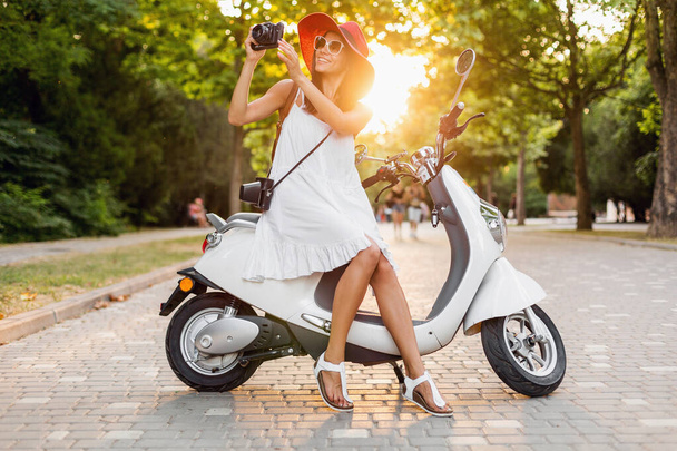 attractive smiling woman riding on motorbike in street in summer style outfit wearing white dress and red hat traveling on vacation, taking pictures on vintage photo camera - Foto, Imagen