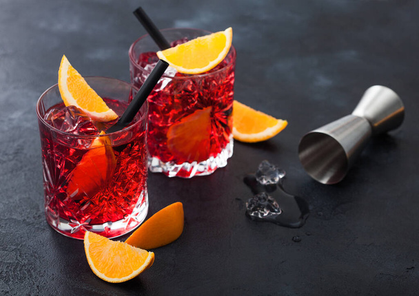 Negroni cocktail in crystal glasses with orange slice and fresh raw oranges with jigger and ice cubes on black background. Top view - Фото, изображение