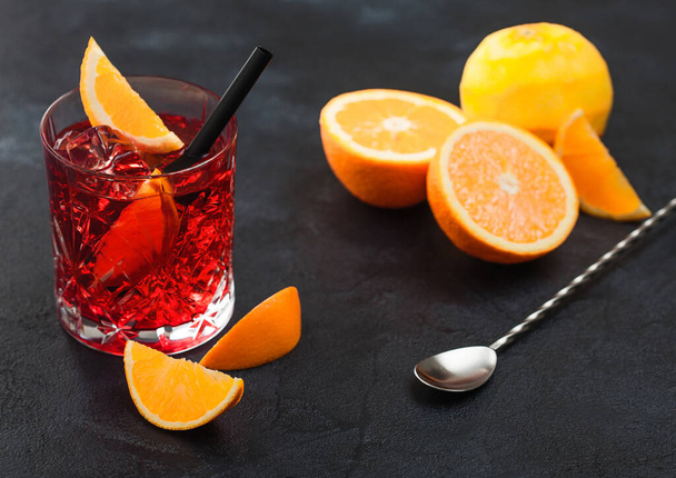 Negroni cocktail in crystal glass with orange slice and fresh raw oranges with spoon on black background. Top view - Fotografie, Obrázek