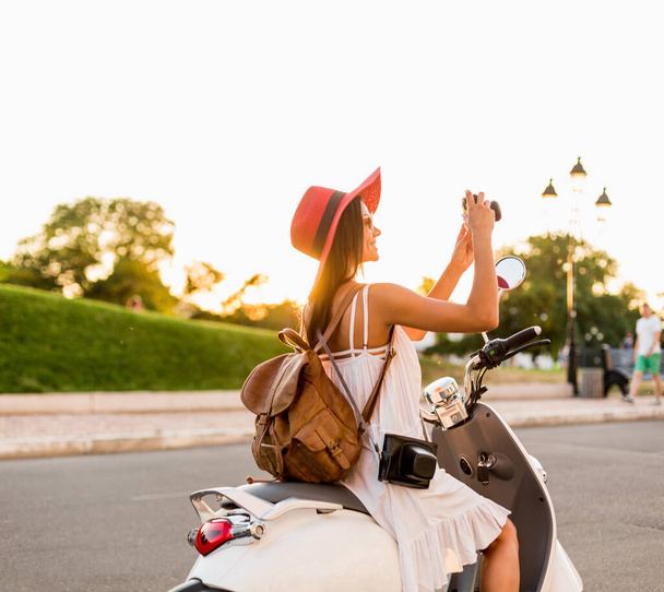 attraente donna sorridente che cavalca in moto in strada in abito stile estivo indossando abito bianco e cappello rosso che viaggia con lo zaino in vacanza, scattando foto sulla macchina fotografica vintage
 - Foto, immagini