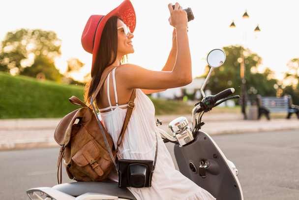 attractive smiling woman riding on motorbike in street in summer style outfit wearing white dress and red hat traveling with backpack on vacation, taking pictures on vintage photo camera - Zdjęcie, obraz