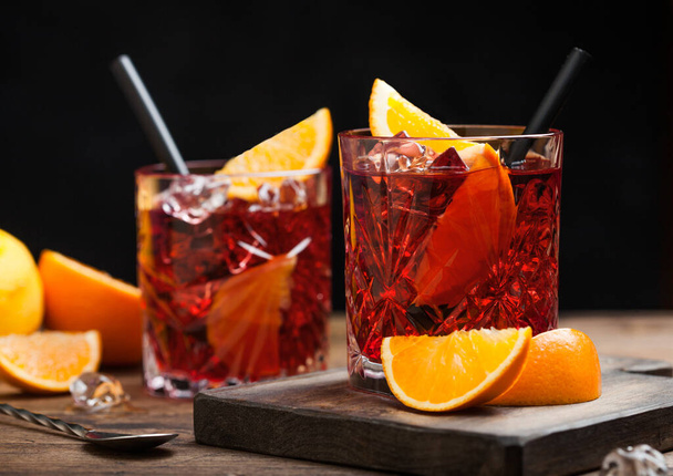 Negroni cocktail in crystal glasses with orange slice and fresh raw oranges on chopping board with strainer on wood background. Macro - Фото, изображение
