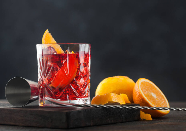 Negroni cocktail in crystal glass with orange slice and fresh raw oranges on chopping board with spoon on wooden background. - Fotoğraf, Görsel