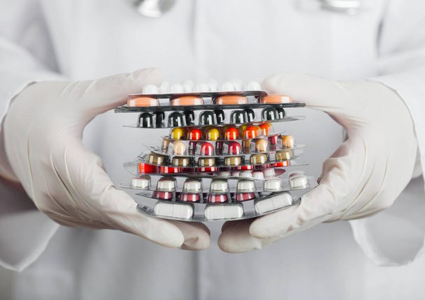 Medical doctor wearing clear latex gloves holding stack of different pills, antibiotics and virus treatment tablets on grey hospital wall. - Фото, зображення