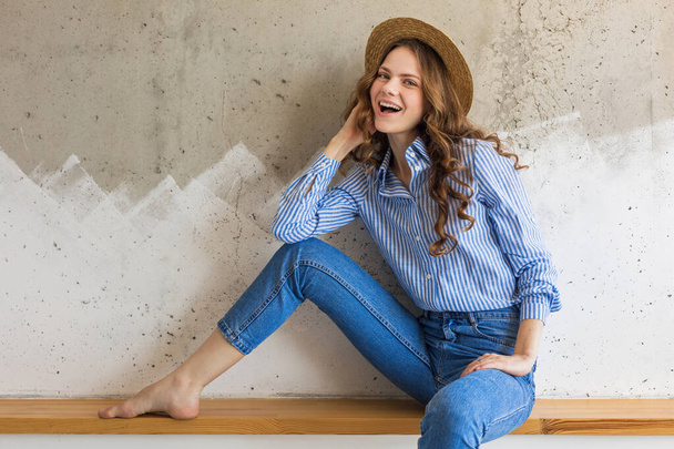 young attractive stylish woman sitting at wall background, straw hat, wearing blue cotton shirt, jeans, casual outfit, hipster style, spring summer fashion trend, smiling face, happy mood, relaxed - Fotoğraf, Görsel