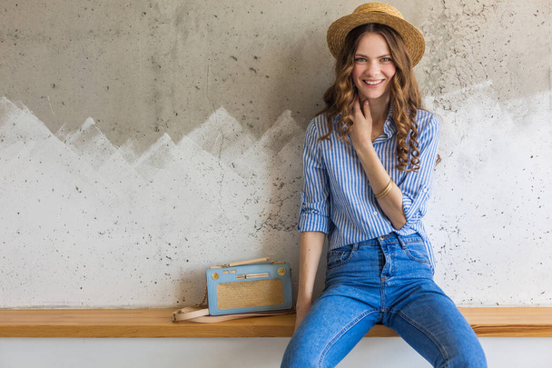 young attractive stylish woman sitting at wall background, holding straw hat, handbag, wearing blue cotton shirt, jeans, casual outfit, hipster vintage style, spring summer fashion trend, accessories - Foto, Imagem