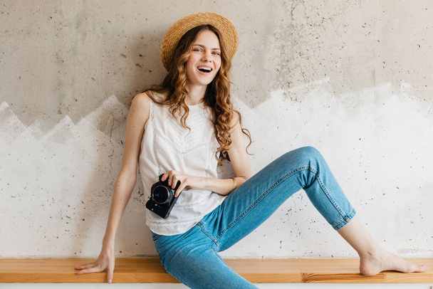 young pretty smiling stylish woman wearing blue denim jeans and white shirt against wall in straw hat holding vintage photo camera, traveler in summer outfit, fashion trend - Photo, image