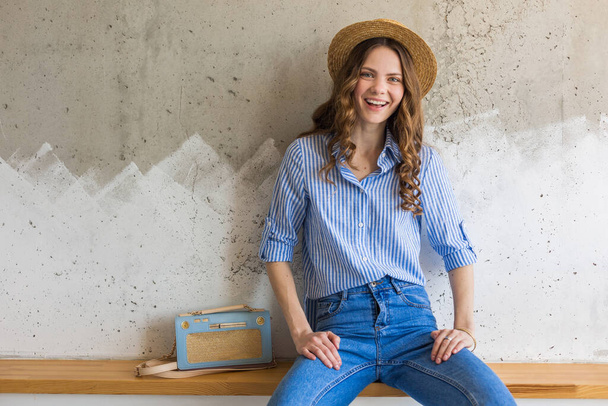 νεαρή ελκυστική κομψή γυναίκα κάθεται σε φόντο τοίχου, ψάθινο καπέλο, φορώντας μπλε βαμβακερό πουκάμισο, τζιν, casual outfit, hipster στυλ, άνοιξη καλοκαίρι μόδα τάση, χαμογελαστό πρόσωπο, χαρούμενη διάθεση, χαλαρή - Φωτογραφία, εικόνα