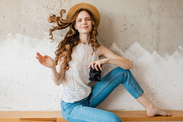 young pretty smiling woman wearing blue denim jeans and white shirt sitting against wall in straw hat holding vintage photo camera, traveler in summer outfit, fashion trend, waving long curly hair - Fotó, kép