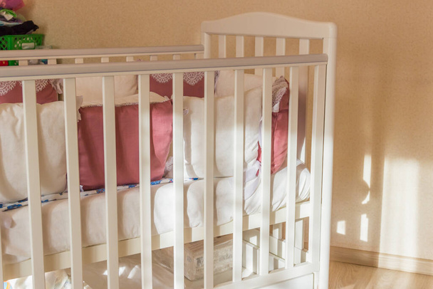 Baby bed with white and Burgundy color pillows with laces. Cot on wheels in the bedroom with pastel yellow colors in the morning. Harsh sun light come from window.Minimalistic interior - Photo, Image