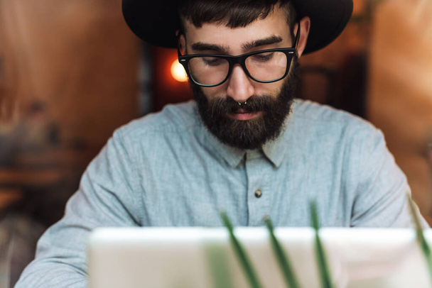 Фото зосередженого молодого чоловіка в окулярах, використовуючи ноутбук, сидячи за столом в кафе в приміщенні
 - Фото, зображення