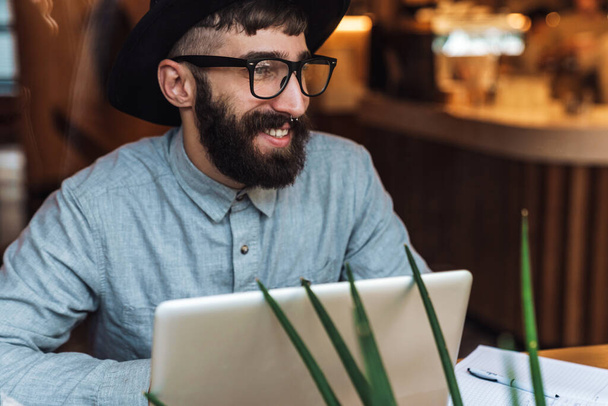 Photo of joyful young man wearing eyeglasses using laptop and smiling while sitting at table in cafe indoors - Fotografie, Obrázek