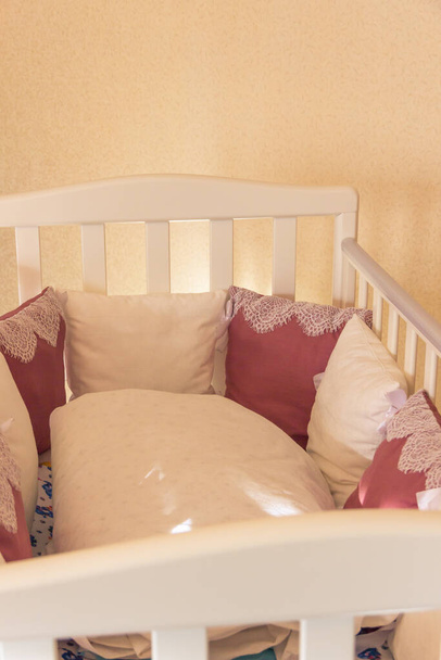 Cama de bebé interior con almohadas de color blanco y burdeos con cordones. Cuna sobre ruedas en el dormitorio con colores amarillos pastel. Interior minimalista
 - Foto, Imagen