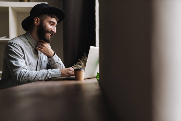 Photo of laughing young man wearing hat typing on laptop and drinking coffee while sitting at table in cafe indoors - Zdjęcie, obraz