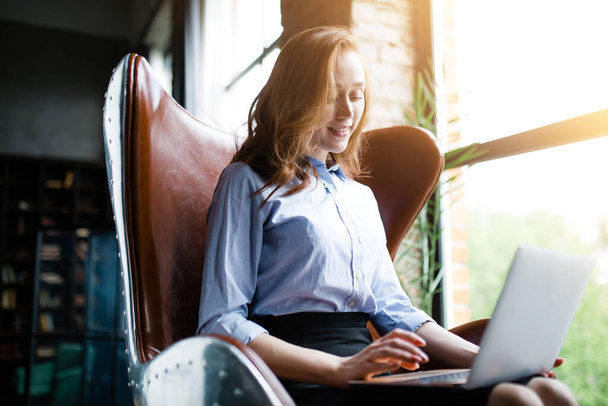 Businesswoman sitting in a leather chair with a laptop on her lap and smiling on the background of a panoramic window. - Foto, Bild