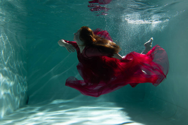 Beautiful girl underwater in a red dress swims in the pool. Tenderness and elegance. Bubbles and a lot of water - Фото, зображення