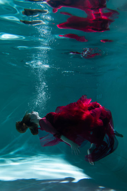 Όμορφη κοπέλα κάτω από το νερό με κόκκινο φόρεμα κολυμπάει στην πισίνα. Ευαισθησία και κομψότητα. Φυσαλίδες και πολύ νερό. - Φωτογραφία, εικόνα