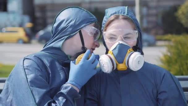 Close-up of young joyful man whispering on ear of pretty woman in chemical suit and respirator. Funny smiling Caucasian couple gossiping outdoors. Covid-19, humor, fun, lifestyle. - Metraje, vídeo