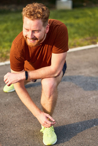 Hombre joven usando reloj de mano durante el ejercicio / estiramiento en el parque urbano
. - Foto, imagen