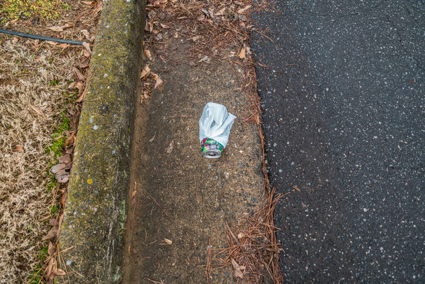 Odrzucony napój może zawinięty w plastikową torbę wyrzuconą na ulicę wzdłuż krawężnika drogi zanieczyszczającej środowisko z powodu niedbalstwa. - Zdjęcie, obraz