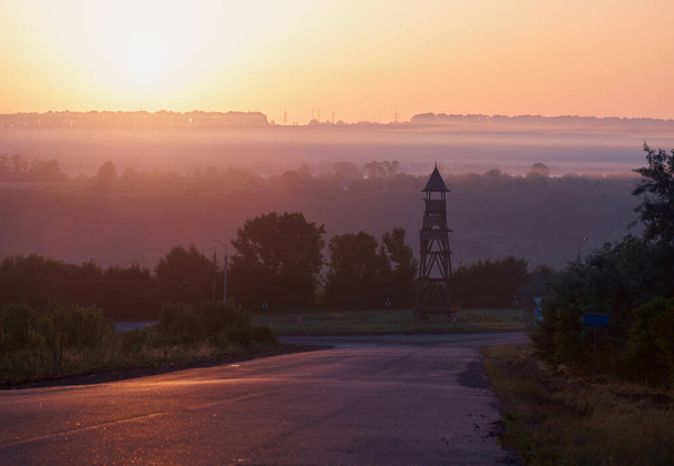 Туманний ранок на сході сонця. Блакитний туман на сонці. Старовинна дерев'яна вежа посеред дорожнього кільця. Асфальтована дорога в тумані. Електричні полюси на горизонті
. - Фото, зображення