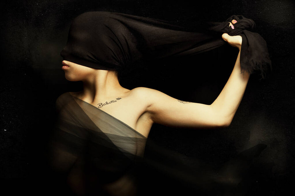 Γυναίκα τυφλωμένη από ένα ύφασμα που κρύβει το πρόσωπο. Εξέγερση και σιωπή. Μια νεαρή γυναίκα τυφλώνεται από ένα ύφασμα τραβηγμένο από το χέρι. Καλλιτεχνικό πορτρέτο με μαύρο φόντο. Ένα μεταξωτό πέπλο που καλύπτει το στήθος. - Φωτογραφία, εικόνα