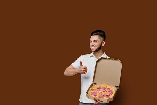 Online pizza safety delivery servise from restaurant. Young caucasian courier man shows thumb up and smile. Pizza with cheese boards advertise. Isolated brown background with empty space for advertise - Photo, Image
