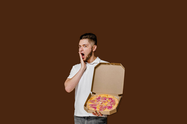 Online pizza biztonsági kézbesítő servise étteremből. A fiatal meglepett futár kinyitotta a száját. Pizza sajtos táblákkal reklámozza. Elszigetelt barna háttér üres helyet hirdetni - Fotó, kép