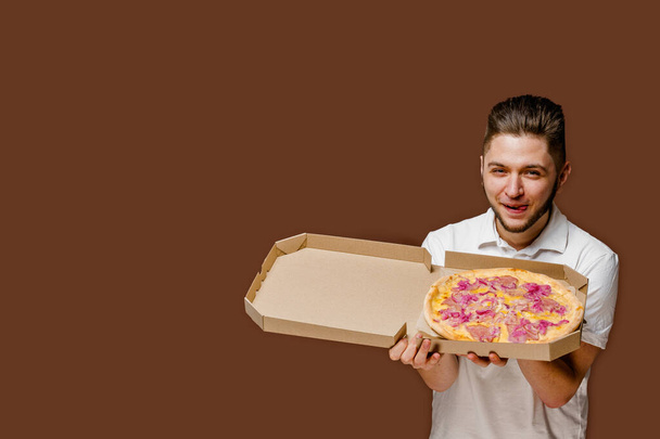 Usługa bezpieczeństwa dostaw pizzy online z restauracji. Młody, atrakcyjny kurier wącha pizzę i liże usta. puste miejsce na reklamę. Pizza z deskami serowymi. Odizolowane brązowe tło - Zdjęcie, obraz