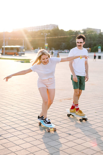 young blondhaired woman longboarding with her partner, boths are in the bright shorts of pink and green colours - Zdjęcie, obraz