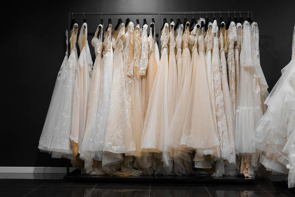 Sukienki ślubne wykonane z jedwabnego szyfonu, tiulu i koronki. Piękna biała kremowa suknia ślubna na wieszakach w salonie weselnym. Perły i kryształy wisiory na rękawach delikatnego koloru sukni ślubnej - Zdjęcie, obraz