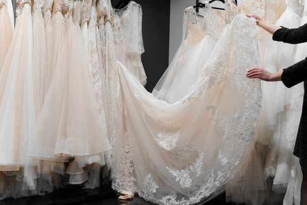 Bride touches luxurious white cream wedding dress on hangers. Dresses made of silk chiffon, tulle and lace. Luxury pearls and crystals pendants on the sleeves of a delicate color of a wedding dress - 写真・画像