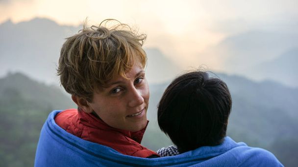 Mladý krásný mezinárodní pár, nejlepší přátelé sdílejí deku. Pozorování východu slunce v horách z pohledu leteckého vrcholu, usmívající se šťastná žena muž, letní dovolená, koncept cestování - Fotografie, Obrázek