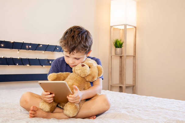 Adorable blond boy sitting on the sofa with teddy bear and reading with tablet pc at home, indoors. Child with tablet computer. Cozy interior. - Photo, Image