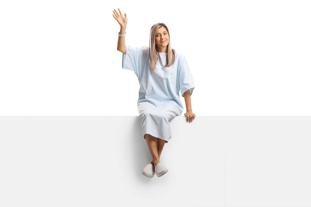 Woman wearing a hospital gown sitting on a blank panel and waving isolated on white background  - Φωτογραφία, εικόνα