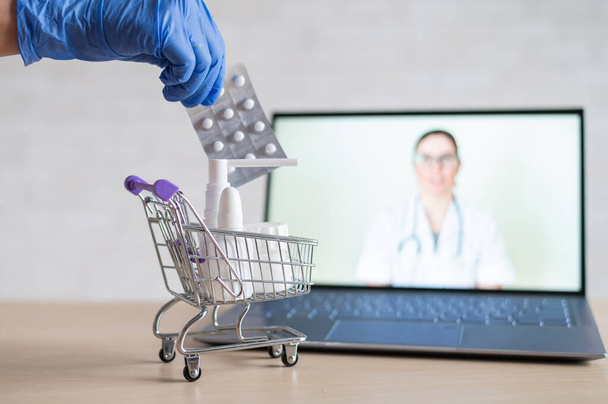 A friendly doctor conducts an online consultation on a laptop. A gloved pharmacist puts medicine into a mini shopping cart. The concept of distance medicine. Order drugs online. - Foto, Imagem