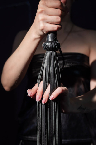 BDSM concept. A woman in a leather dress holds a black whip with a long fringe. Role-playing games for adults. Close-up of sex toys for domination. - Φωτογραφία, εικόνα