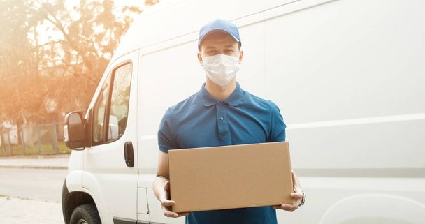 Portrait of young courier in mask standing near delivery car and holding carton box - Фото, изображение