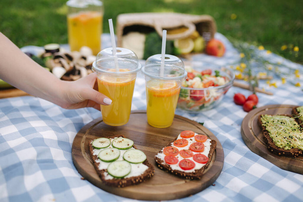 Mujer tomar jugo de naranja fresca en el picnic vegano al aire libre, Picnic en manta a cuadros azules en el parque. Concepto de comida vegana
 - Foto, Imagen