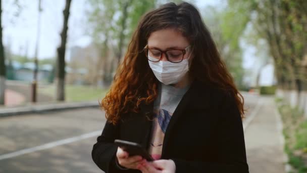 Młoda dziewczyna w masce medycznej używa smartfona w parku. 4k - Materiał filmowy, wideo