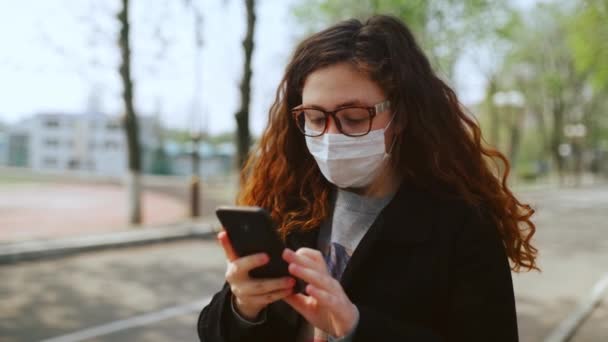 Młoda dziewczyna w masce medycznej używa smartfona w parku. 4k - Materiał filmowy, wideo