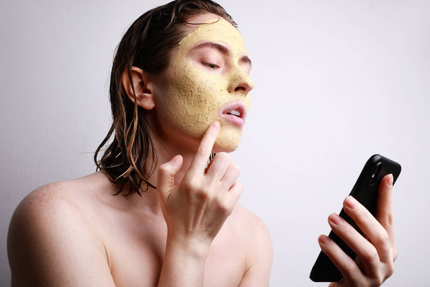 Close-up portrait of young caucasian woman with facial clay mask using her smartphone watching videos. Space for text. - Photo, image