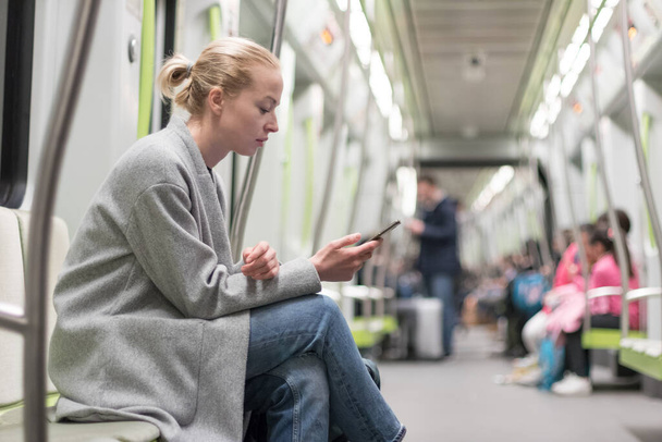 Portret uroczej dziewczyny wpisującej wiadomość na telefonie komórkowym w prawie pustym publicznym pociągu metra. Pozostawanie w domu i dystansowanie społeczne zalecane z powodu wybuchu pandemii wirusa korony - Zdjęcie, obraz