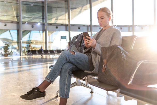 Alkalmi szőke fiatal nő használja a mobilját, miközben arra vár, hogy felszálljon egy repülőre a reptér indulási kapujánál. Koronavírus-világjárvány miatt kiürült repülőtéri terminál - Fotó, kép