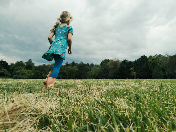かわいい女の子が草原を走る。雨のブーツの子供の女の子の足。自由の無実と青少年の概念。子供のための夏の楽しい屋外活動。後ろから見るんだ。低角度表示.  - 写真・画像