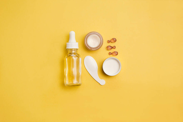 Skin rejuvenation concept - glass face serum bottle, eye cream, face oil capsules on yellow background. - Photo, Image