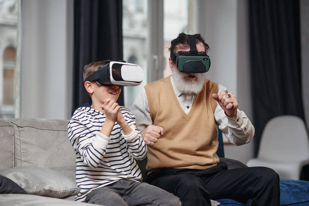 Portrait of excited senior man using VR glasses sitting on sofa at home with laughing grandson beside him - Zdjęcie, obraz