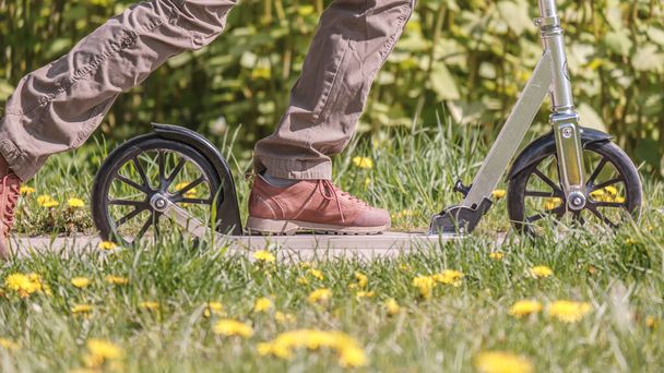 Dişi bacaklar baharda, çiçek açan çimenlerin arasında kaldırımda scooter kullanıyor.. - Fotoğraf, Görsel