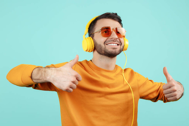 Gleeful bearded young man wearing sunglasses listening to music showing ok gesture on cyan background. - Foto, Imagem